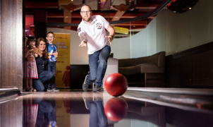 man smiling and bowling with a red ball whilst family look on in the background