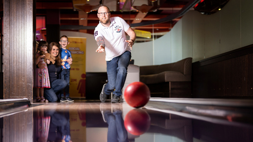 man smiling and bowling with a red ball whilst family look on in the background